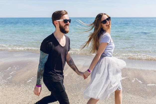 Handsome guy in black sunglasses with beard walks on the beach near sea holding a hand of pretty woman with long hair