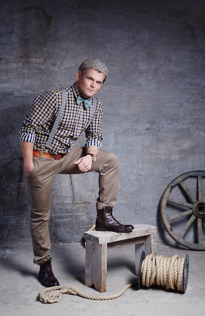 Free photo handsome guy in checkered shirt with blue bow tie posing near gre concrete wall with wooden wheel.