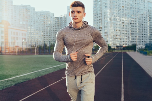 Handsome guy in gray sport suit  is running at workout in the morning on stadium. He is listening to music through headphones and looking to side.