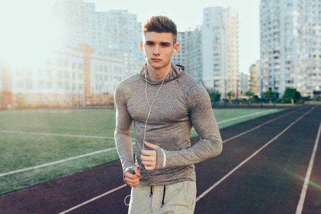 Handsome guy in gray sport suit is running at workout in the morning on track on city background. He is listening to music through headphones and looking to camera.