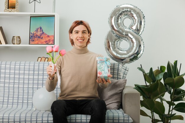 handsome guy on happy women day holding flowers with present sitting on sofa in living room