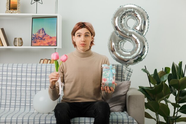 Handsome guy on happy women day holding flowers with present sitting on sofa in living room