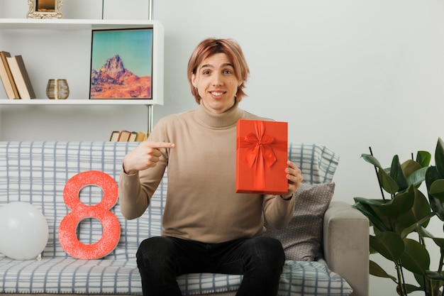 Handsome guy on happy women day holding and points at present sitting on sofa in living room