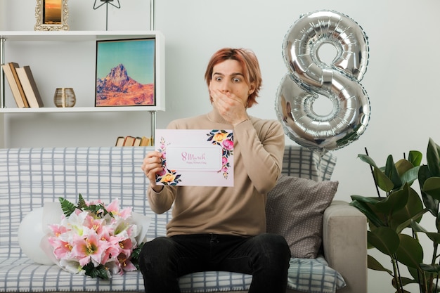 Handsome guy on happy women day holding postcard sitting on sofa in living room