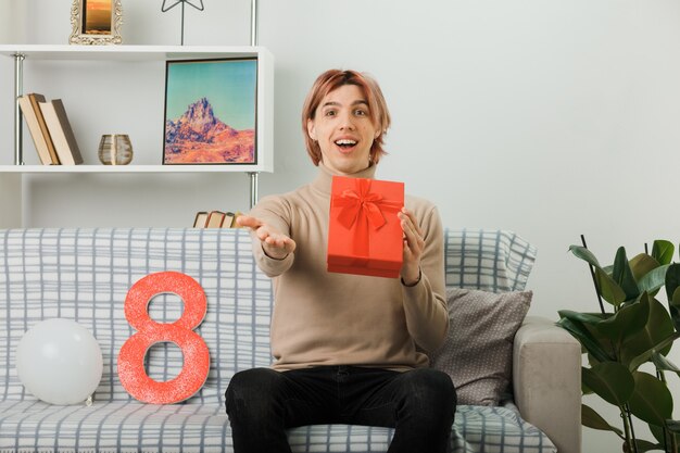 Bel ragazzo in una felice giornata delle donne con un regalo seduto sul divano in soggiorno