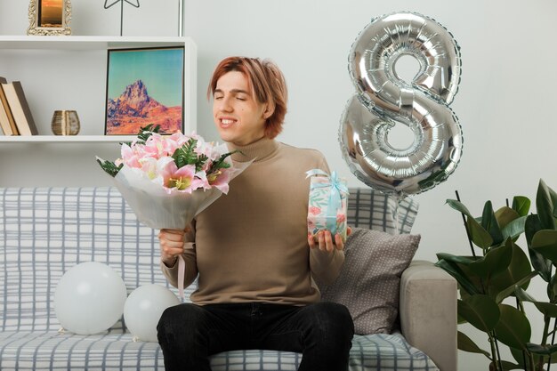 Handsome guy on happy women day holding present with bouquet sitting on sofa in living room