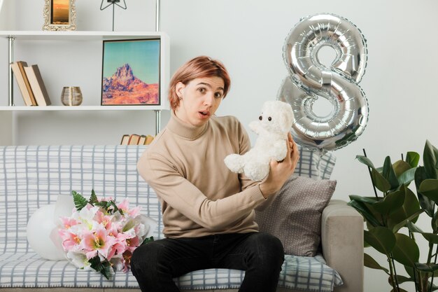 Handsome guy on happy women day holding teddy bear sitting on sofa in living room