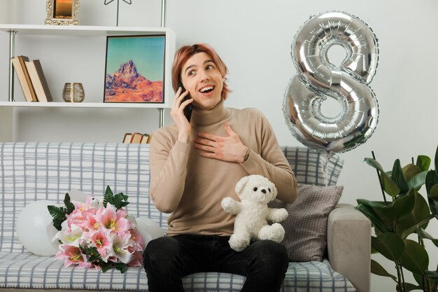 Handsome guy on happy women day holding teddy bear speaks on phone sitting on sofa in living room