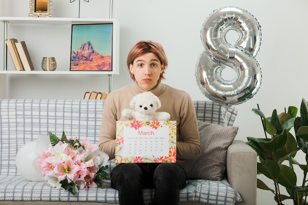 Handsome guy on happy women day holding teddy bear with calendar sitting on sofa in living room