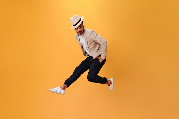 Handsome guy in hat and suit is jumping high on isolated background Young man in beige jacket and white shirt dancing on orange backdrop