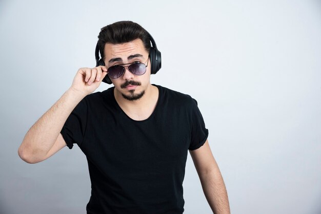 handsome guy model in headphones posing with sunglasses against white wall . 