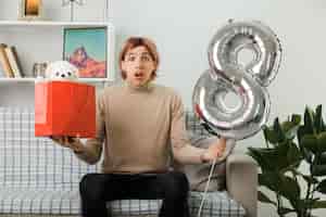 Бесплатное фото Красивый парень в счастливый женский день держит воздушный шар номер восемь с подарочной сумкой, сидя на диване в гостиной