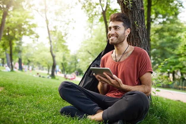 ハンサムな男が公園の芝生に座って、デジタルタブレットの本を読んで、wifiを接続し、ソーシャルメディアを見る