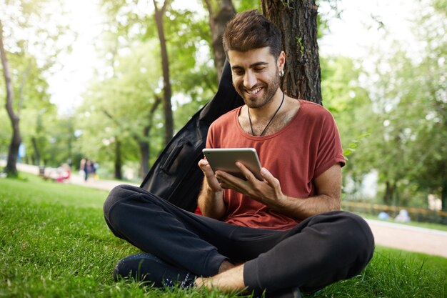 Красивый парень сидит в траве в парке, читает книгу на цифровом планшете, подключает Wi-Fi и просматривает социальные сети
