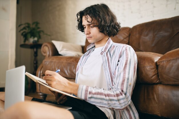 Handsome guy sitting on floor making notes while listening to educational course, studying online. Serious guy working from home