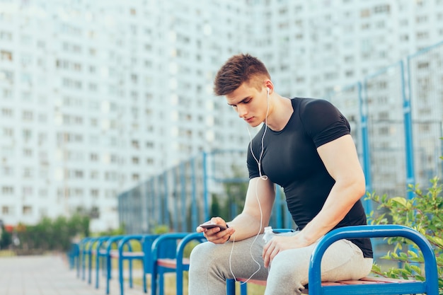 スポーツブラックのtシャツとグレーのスポーツパンツでハンサムな男は、都市とスタジアムの背景にベンチに座っています。彼は電話でタイプし、ヘッドフォンで音楽を聴いています。