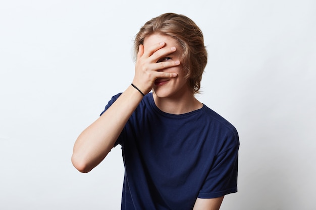 Handsome guy with attractive appearance, hiding his face with hand, looking through fingers, having shy expression. Young hipster guy not wanting to be photographed, covering his face with hand