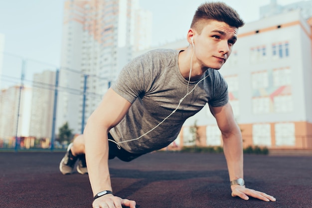 Handsome guy with a good body push-ups in the morning on stadium. He wears sport clothes, listening to music through headphones. He looks tense.