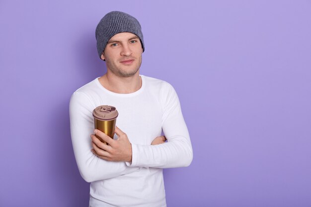 캐주얼 흰색 긴 소매 셔츠와 회색 모자를 입고, 테이크 아웃 커피를 손에 들고, 기쁘게 표현 잘 생긴 남자는 라일락 벽 위에 절연 뜨거운 음료를 즐긴다