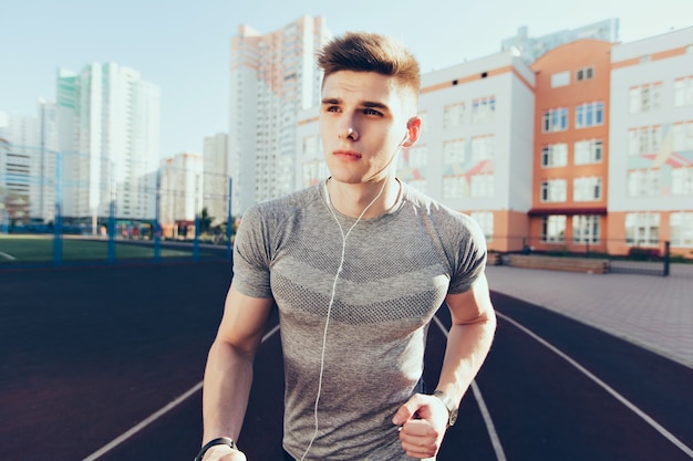 Handsome guy at workout running in the morning on stadium. He wears gray T-shirt, listening to music through headphones.