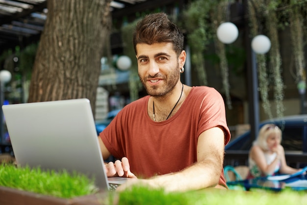 Красивый счастливый бородатый мужчина в наушниках, используя ноутбук в летнем кафе, весело улыбаясь