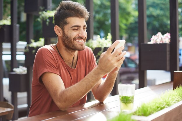 Handsome happy guy sitting in cafe, drinking lemonade and using mobile phone, texting message