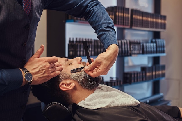 A handsome hipster bearded male sitting in an armchair in a barber shop while hairdresser shaves his beard with a dangerous razor.