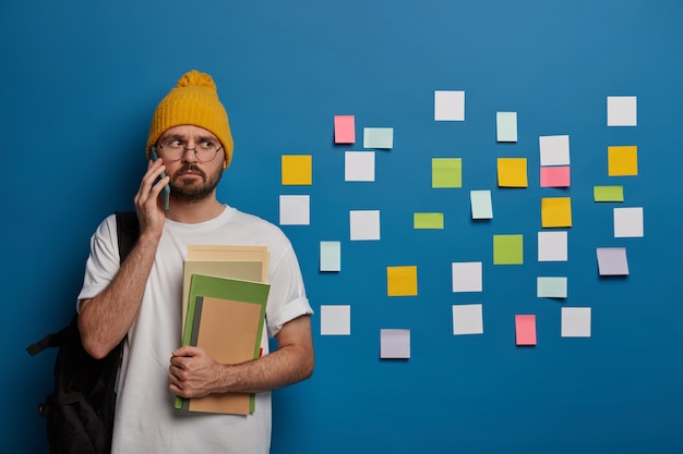 Handsome hipster calls friend after lectures, returns from college, stands with textbooks and rucksack