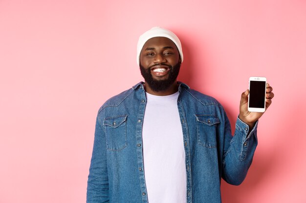 Handsome hipster guy in beanie and denim shirt smiling, showing mobile phone screen with happy face, introduce application, standing over pink background