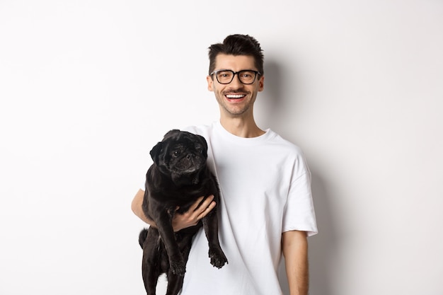 Handsome hipster guy holding his funny black pug dog, smiling at camera, standing over white background.