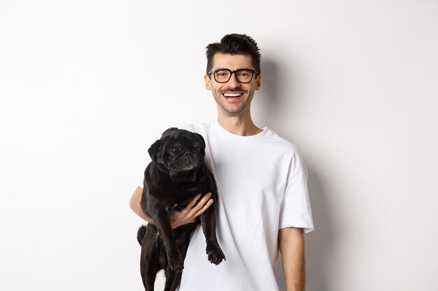 彼の面白い黒いパグ犬を保持し、カメラに微笑んで、白い背景の上に立っているハンサムな流行に敏感な男。