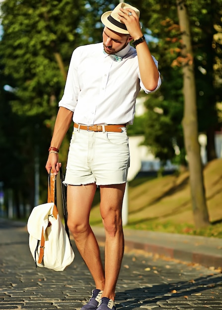 バッグと帽子でポーズをとってスタイリッシュな夏服でハンサムなヒップスターモデル男