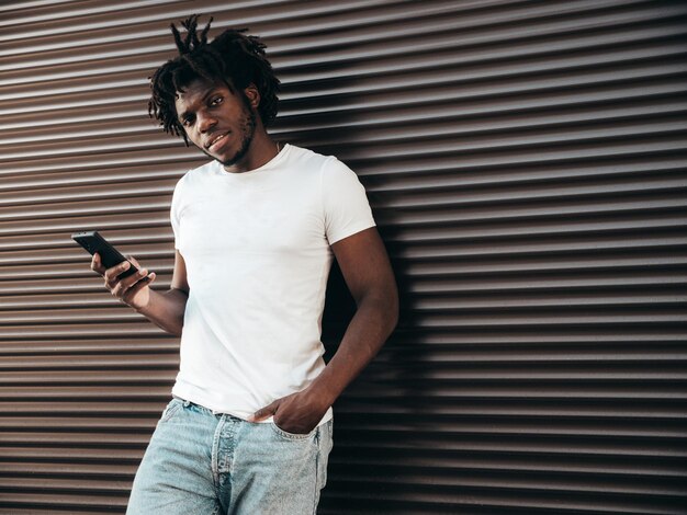 잘생긴 힙스터 모델흰색 여름 티셔츠를 입은 형태가 없는 아프리카 남자 향취 헤어스타일을 한 패션 남성거리에서 포즈를 취함휴대폰 앱을 사용하여 스마트폰 화면을 바라보고 있습니다.