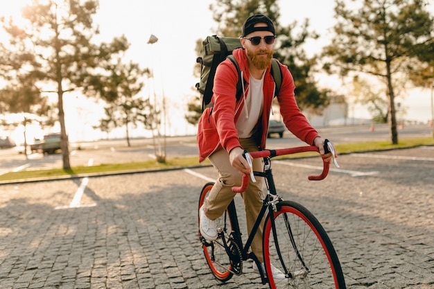 Красивый бородатый мужчина в стиле хипстера в красной толстовке с капюшоном и солнцезащитных очках, едущий в одиночестве с рюкзаком на велосипеде, здоровый активный образ жизни, путешественник, рюкзаком