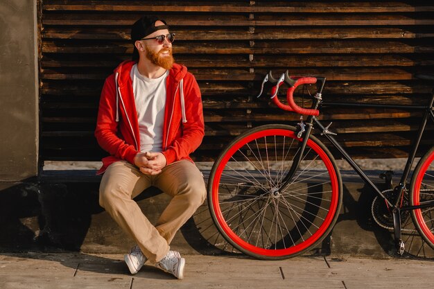 Handsome hipster style bearded man in red hoodie sitting relaxing alone with backpack and bicycle in morning sunrise by the sea healthy active lifestyle traveler backpacker