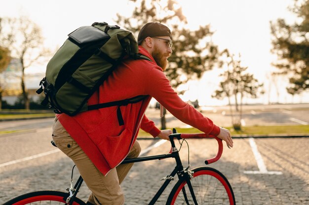 赤いパーカーとサングラスのハンサムなヒップスタースタイルのひげを生やした男は、自転車の健康的なアクティブなライフスタイルの旅行者のバックパッカーにバックパックと一緒に乗っています