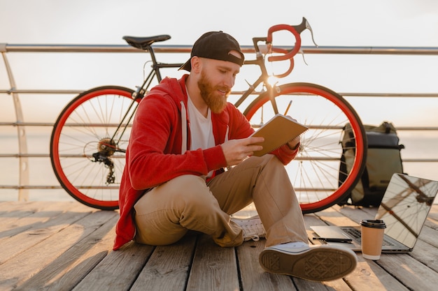 海の健康的なアクティブなライフスタイルの旅行者のバックパッカーによる朝の日の出でバックパックと自転車でメモを書くオンラインフリーランサーを勉強しているハンサムなヒップスタースタイルのひげを生やした男