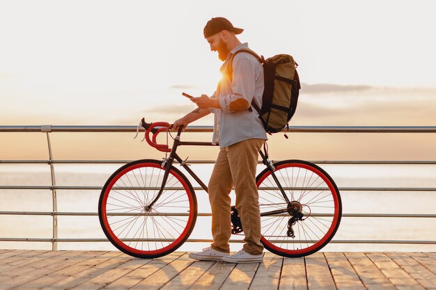 Handsome hipster style bearded man traveling with backpack on bicycle using phone in morning sunrise by the sea, healthy active lifestyle