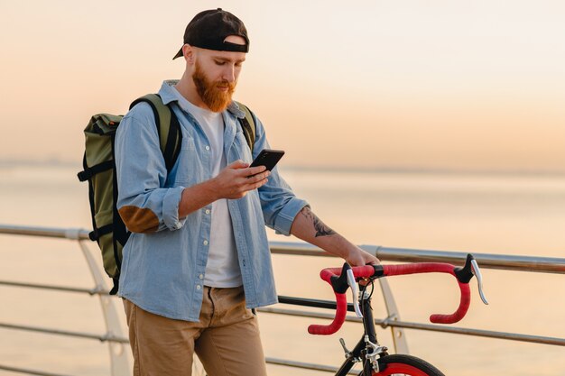 Handsome hipster style bearded man using phone traveling with backpack and bicycle in morning sunrise by the sea, healthy active lifestyle traveler backpacker