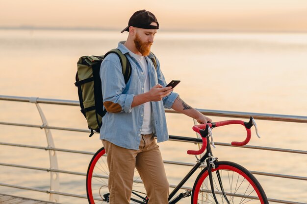 Handsome hipster style bearded man with backpack wearing denim shirt and cap traveling with bicycle in morning sunrise by the sea drinking coffee, healthy active lifestyle traveler backpacker