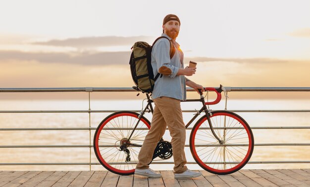 Красивый бородатый мужчина в стиле хипстера с рюкзаком в джинсовой рубашке и кепке с велосипедом на утреннем восходе солнца у моря, пьющий кофе, путешественник, путешествующий по здоровому активному образу жизни