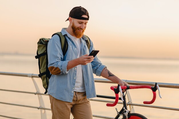 Handsome hipster style bearded man with backpack wearing denim shirt and cap with bicycle in morning sunrise by the sea drinking coffee, healthy active lifestyle traveler backpacker