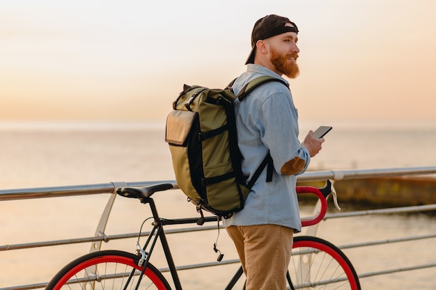 Красивый бородатый мужчина в стиле хипстера с рюкзаком в джинсовой рубашке и кепке с велосипедом на утреннем восходе солнца у моря, пьющий кофе, путешественник, путешествующий по здоровому активному образу жизни