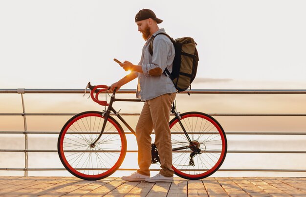 Handsome hipster style bearded man with backpack wearing denim shirt and cap with bicycle in morning sunrise by the sea drinking coffee, healthy active lifestyle traveler backpacker