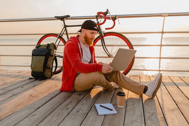 Красивый бородатый мужчина в стиле хипстера, работающий онлайн-фрилансером на ноутбуке с рюкзаком и велосипедом в утреннем восходе солнца у моря, здоровый активный образ жизни, путешественник с рюкзаком