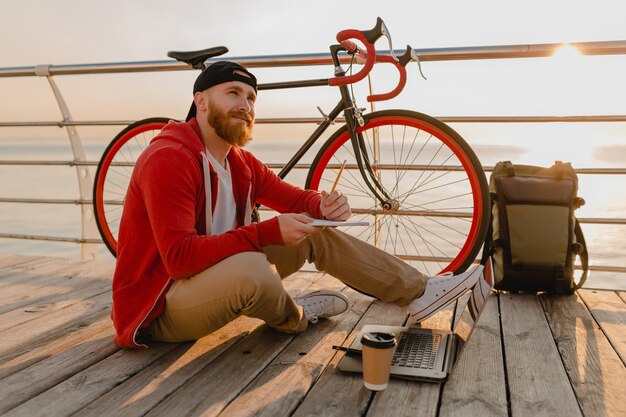 Handsome hipster style bearded man working online freelancer on laptop with backpack and bicycle in morning sunrise by the sea healthy active lifestyle traveler backpacker