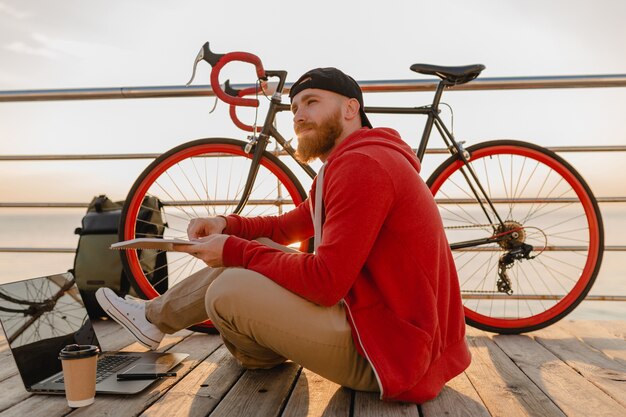 Красивый бородатый мужчина в стиле хипстера, работающий онлайн-фрилансером на ноутбуке с рюкзаком и велосипедом в утреннем восходе солнца у моря, здоровый активный образ жизни, путешественник с рюкзаком