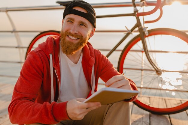 Handsome hipster style bearded man working online freelancer on laptop with backpack and bicycle in morning sunrise by the sea healthy active lifestyle traveler backpacker