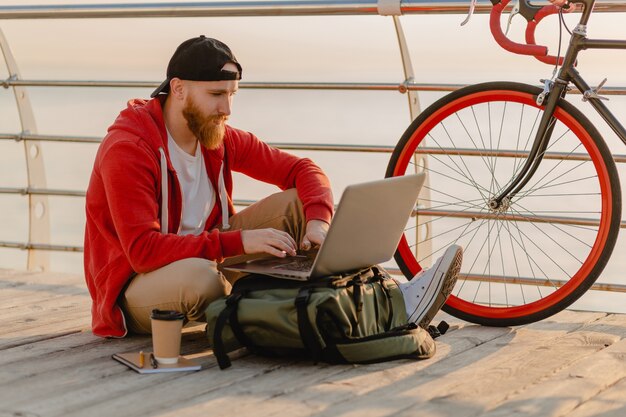 Handsome hipster style bearded man working online freelancer on laptop with backpack and bicycle in morning sunrise by the sea healthy active lifestyle traveler backpacker