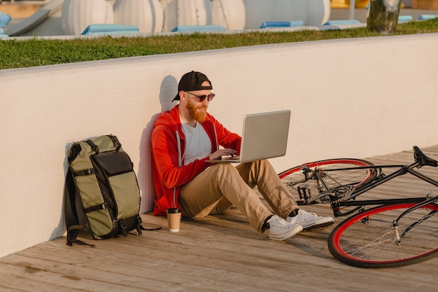 Uomo barbuto bello stile hipster che lavora libero professionista online su laptop con zaino e bicicletta in alba di mattina in riva al mare viaggiatore zaino in spalla sano stile di vita attivo